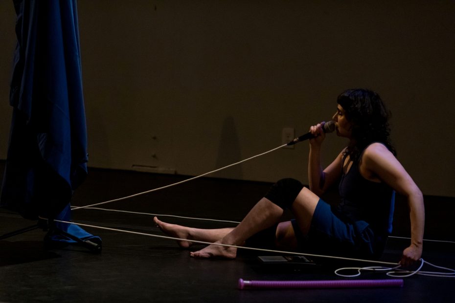 Allie sitting on the floor with a microphone wearing blue against a dark background