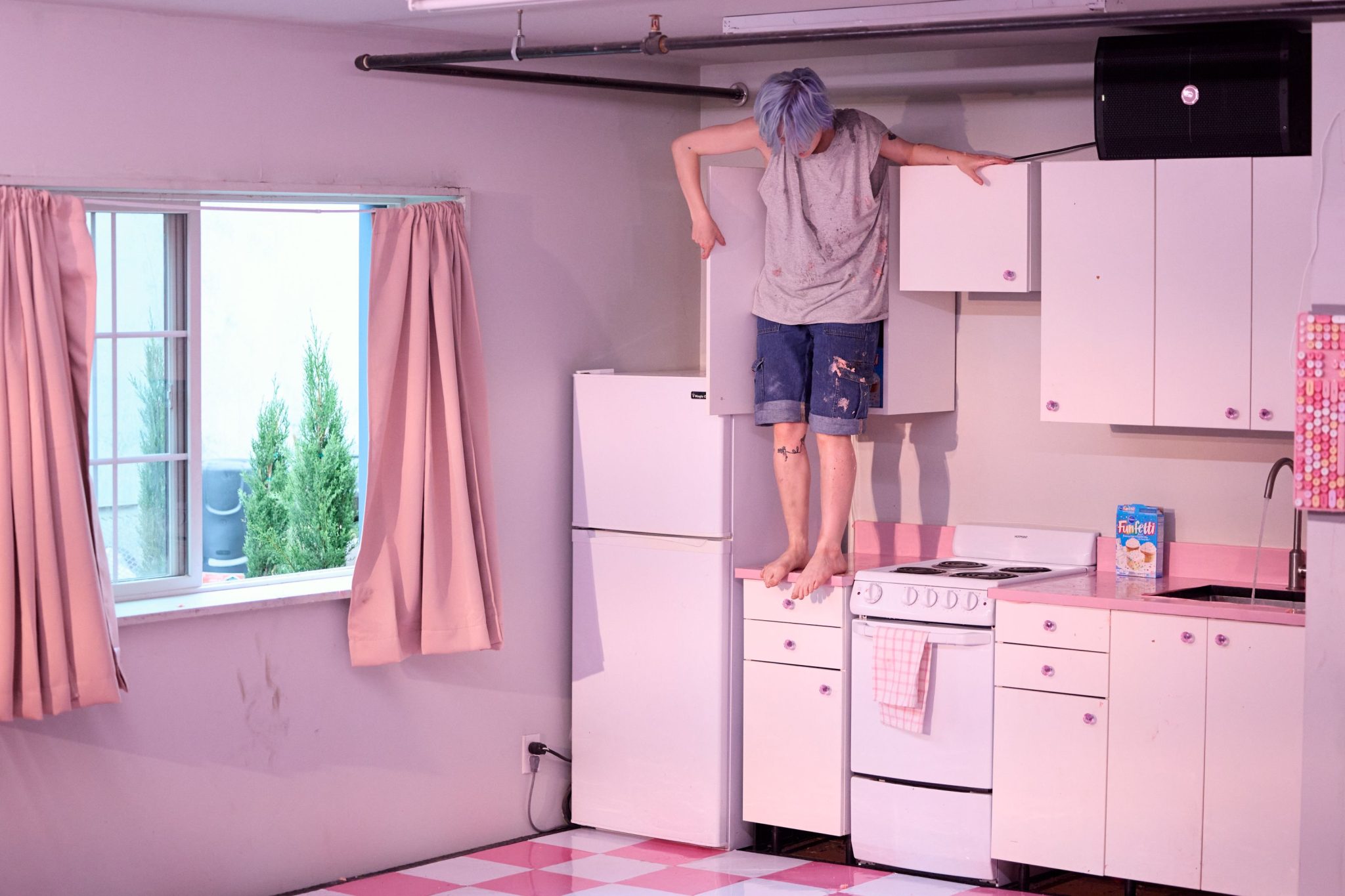 kelly standing on a white counter in a pink a white kitchen wearing a grey tank top and jean shorts