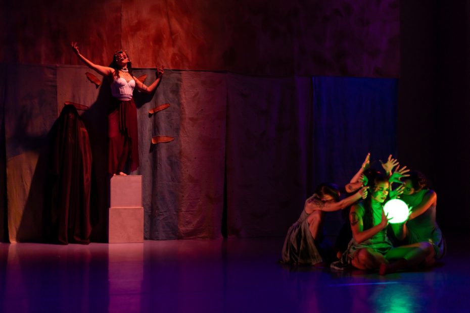 Dancer standing on pedestal with the rest of cast kneeling on floor against a dark background